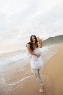 A woman walking along the beach with ocean waves in the background, wearing a white dress.