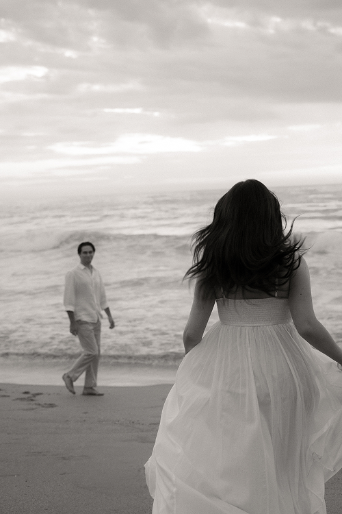 A woman in a white dress standing on a beach with her back to the camera, facing the ocean, while a man in light-colored clothing walks towards her in the background under a cloudy sky.