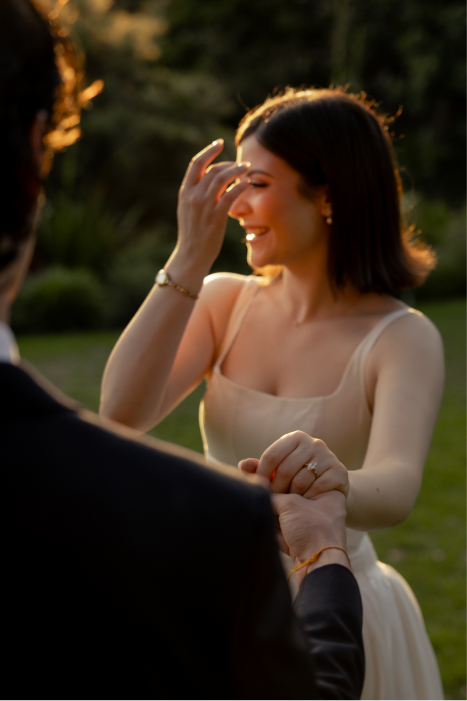 A woman in a cream-colored dress smiling and touching her forehead while holding hands with someone outdoors during sunset.