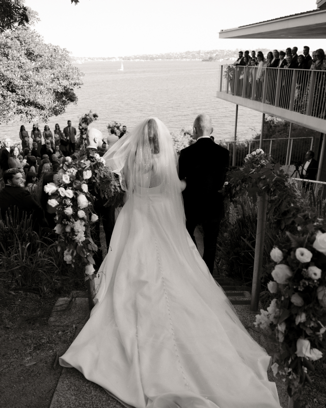 Bride in a wedding gown with a long train and veil, walking down an outdoor aisle with a groom dressed in a suit, accompanied by guests and floral arrangements, near a body of water.