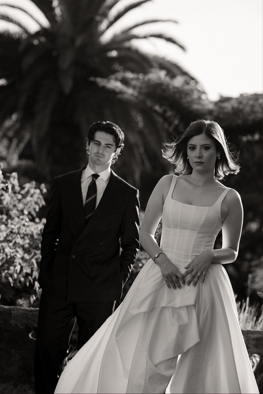 Black and white photo of a woman in a wedding dress standing in front of a man in a suit with a background of trees, possibly at a park or garden.