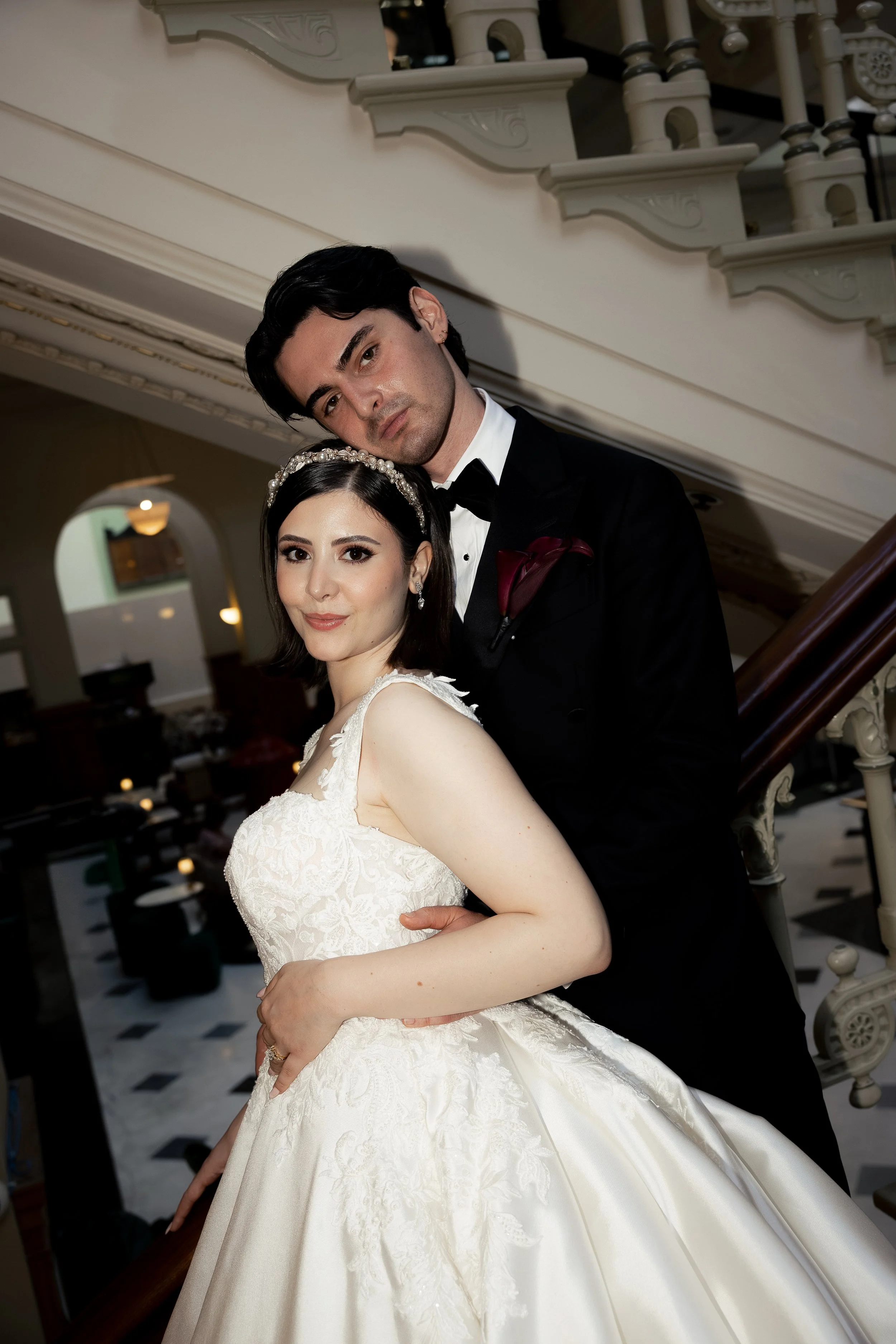 A bride and groom standing on a staircase inside a building. The bride is wearing a white wedding dress with lace details and a pearl headband, and the groom is dressed in a black tuxedo with a bow tie and a red pocket square. The groom is resting his head on the bride's head, and they both are looking at the camera. The background features ornate architectural details and checkered flooring.