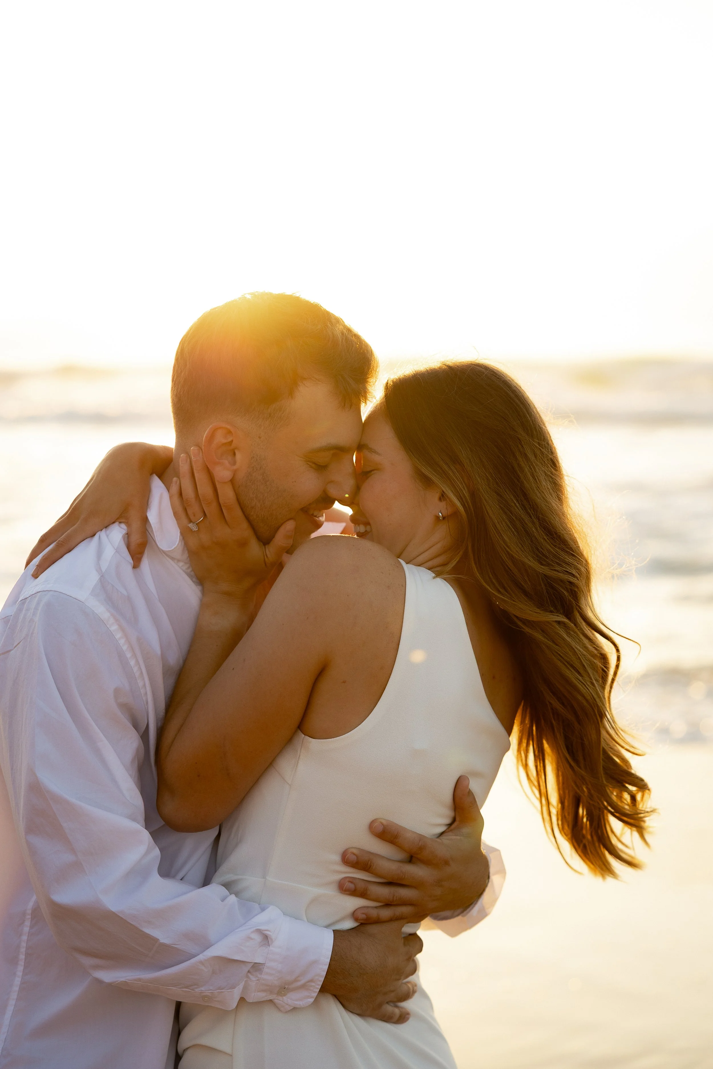 A couple embrace, the man higs his Fiance tighly around her back and she has her hands around his shoulder and one on his face, they are smiling at each other with their foreheads together. The sun shines behind their heads as they stand on the beach