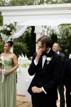 A groom in a tuxedo reacting emotionally during a wedding ceremony outdoors with a floral arch and bridesmaids in green dresses.