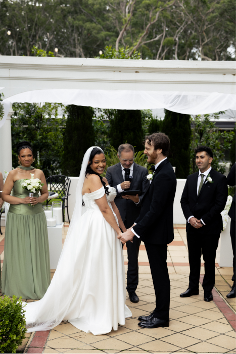 Bride and groom holding hands and smiling during their wedding ceremony outdoors, surrounded by bridesmaid and groomsmen, with officiant reading vows.