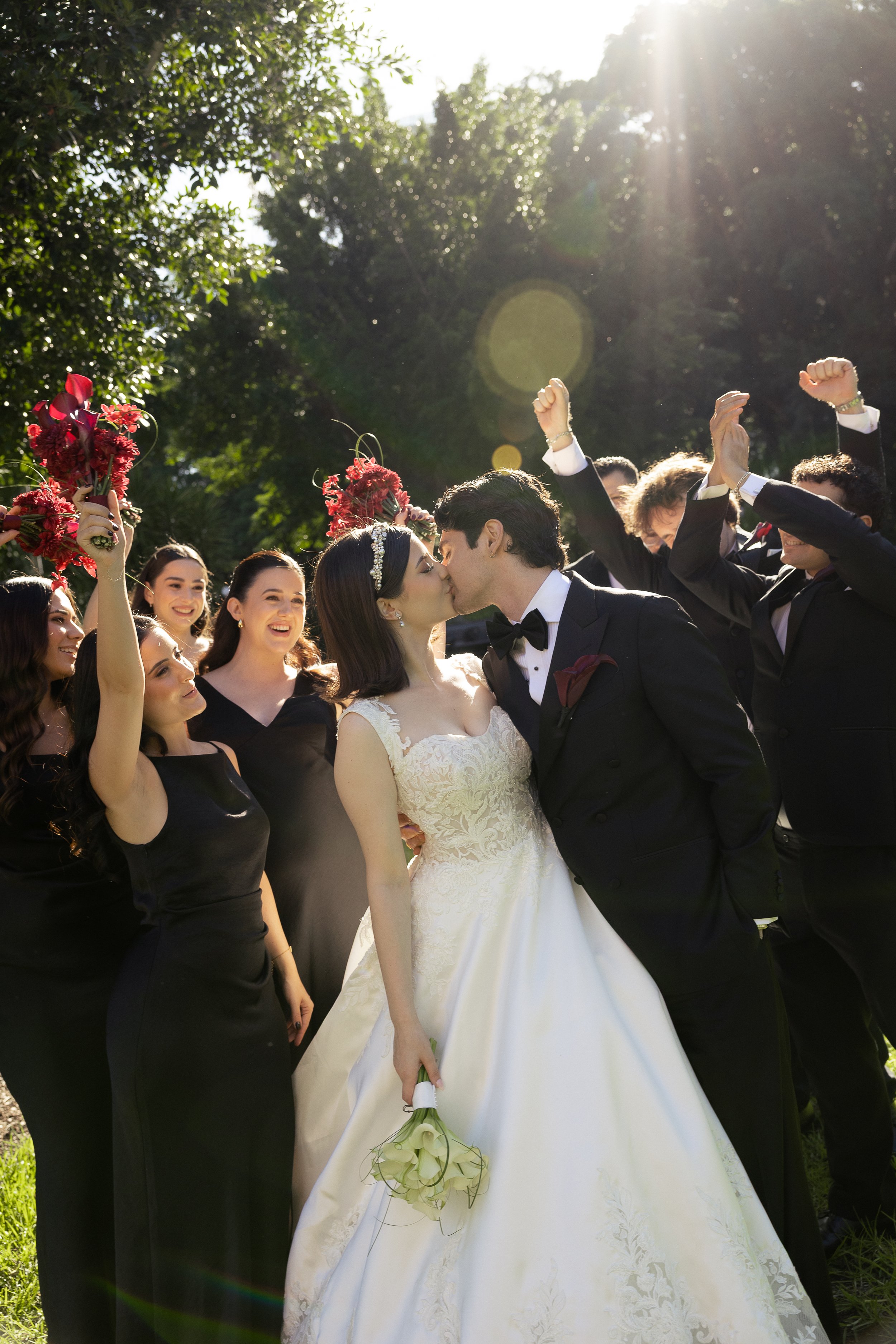 A newlywed couple in wedding attire celebrating outdoors amidst lush greenery, with the bride releasing a burst of confetti or flower petals in the air, and sunlight filtering through trees.