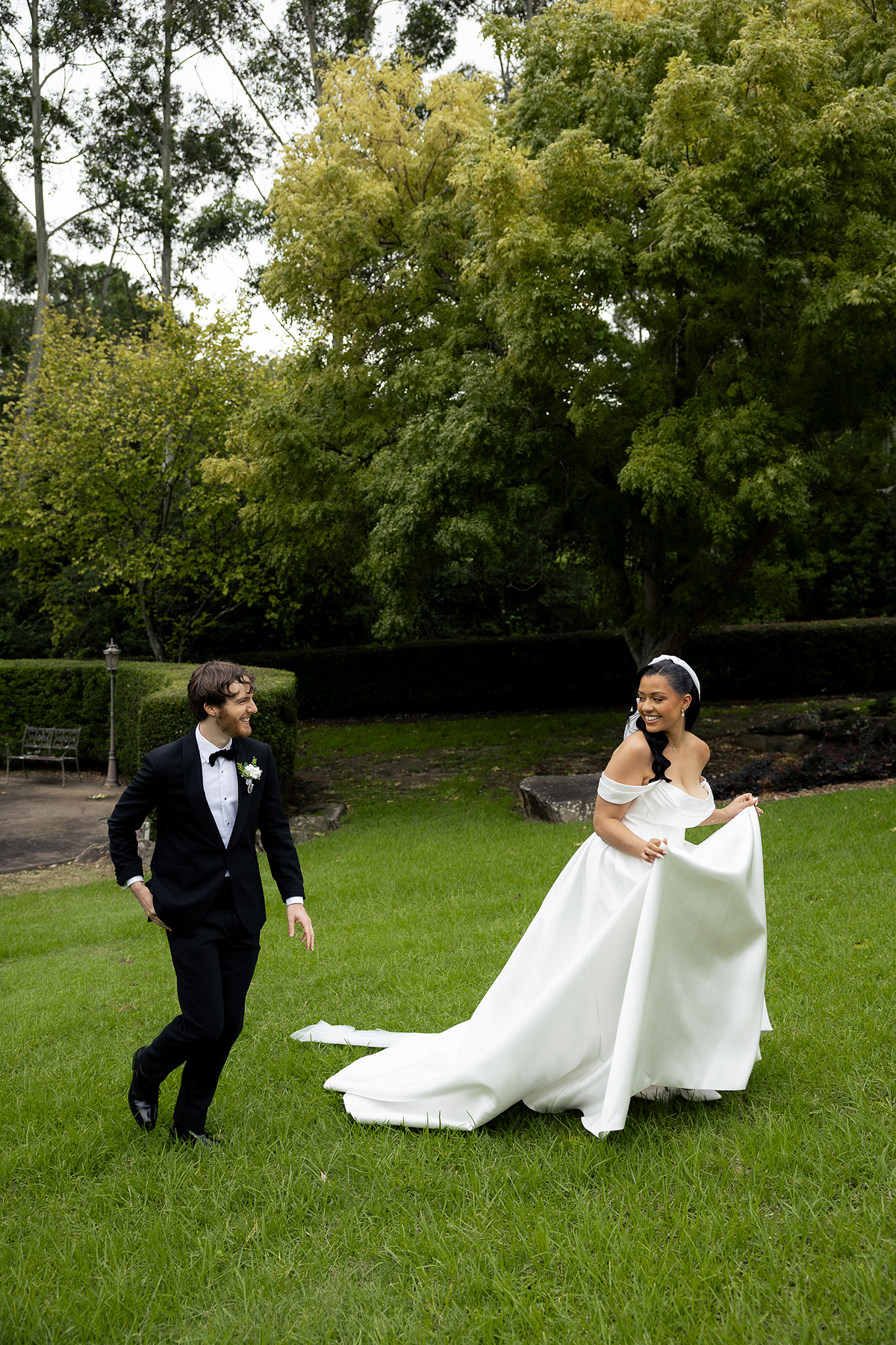 A bride in a white off-shoulder wedding gown with a train and a groom in a black tuxedo with a bow tie, smiling and walking on a grassy area outdoors with trees in the background.