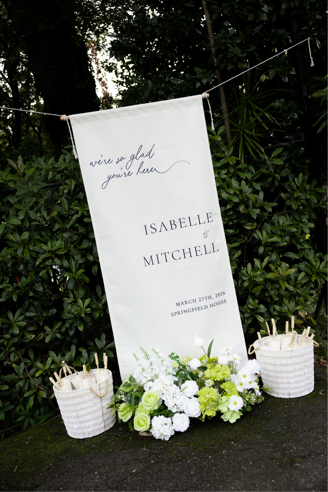 A wedding sign with the names Isabel and Mitchell, date March 27th, 2026, at Springfield House, decorated with white and green flowers and two white woven baskets with small white umbrellas.