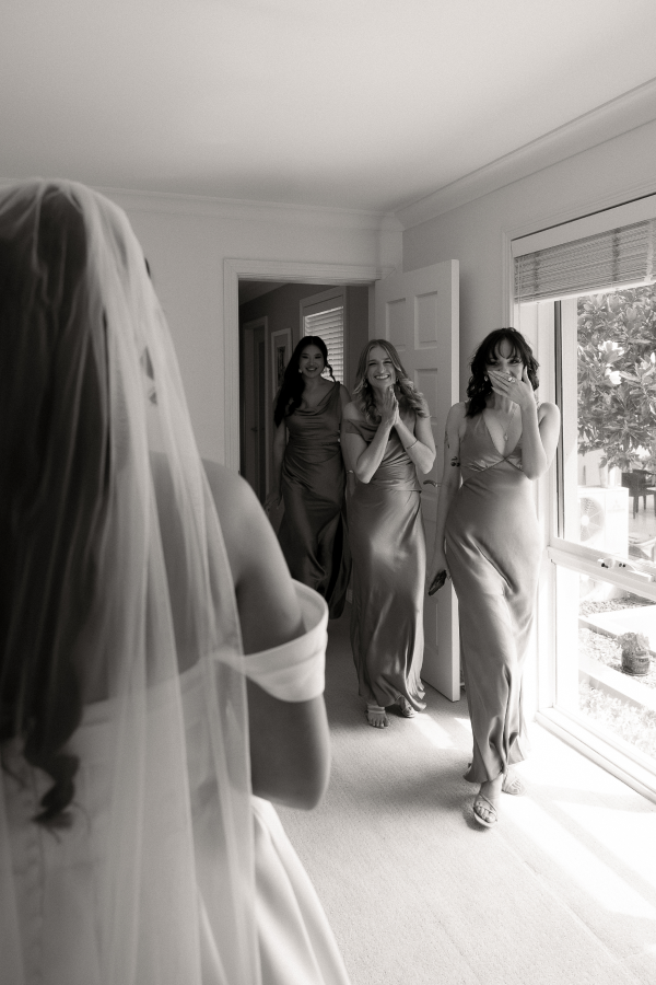 A bride in a wedding dress and veil sees three bridesmaids in satin dresses reacting happily in a sunlit room.