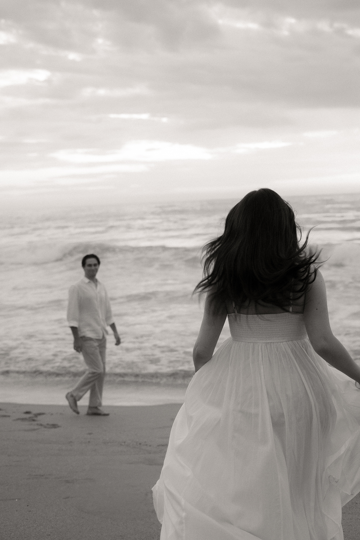 A woman in a flowing dress on a beach with a man in the distance, both looking at each other against the backdrop of the ocean and cloudy sky.