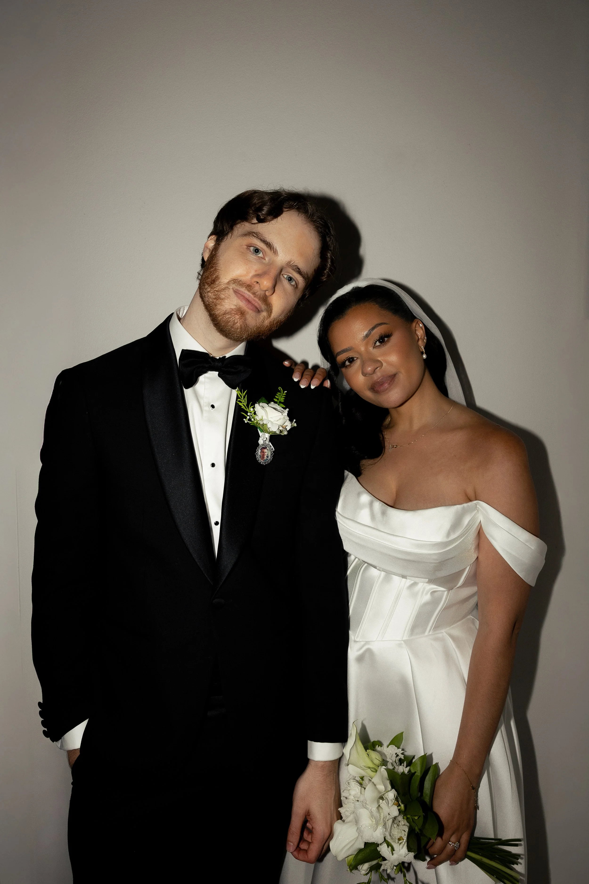 A bride and groom pose closely together at their wedding, the groom wearing a black tuxedo with a bow tie and white shirt, and the bride in an off-shoulder white dress with a veil, holding a bouquet of white flowers.