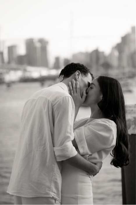 A couple kissing near a city waterfront in black and white.