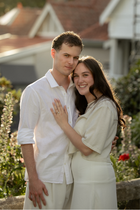 A young couple standing close together outdoors, smiling, with a house and flowers in the background.
