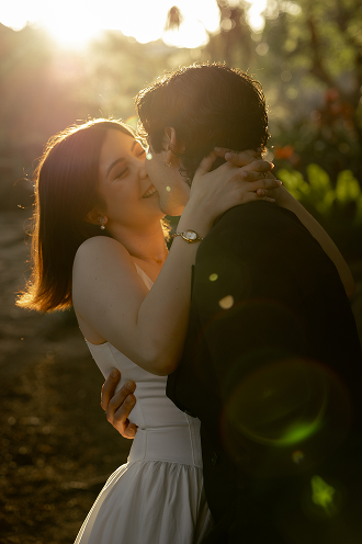 A couple embracing and about to kiss outdoors during sunset, with trees and sunlight in the background.