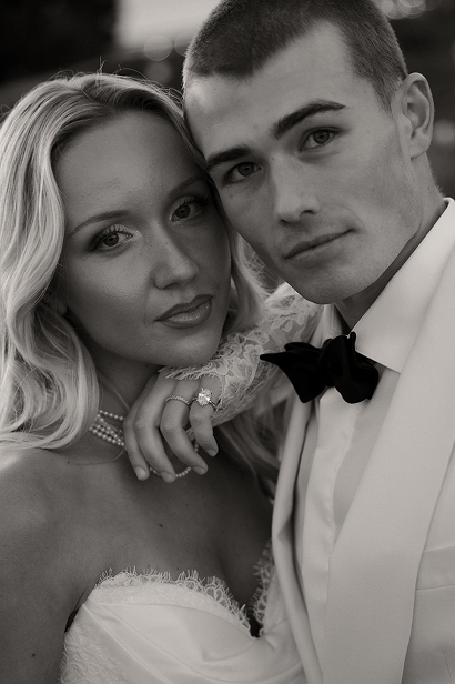 A black and white close-up of a couple dressed in wedding attire, with the woman wearing a lace dress and pearl necklace, and the man in a tuxedo with a bow tie, both looking at the camera with serious expressions.