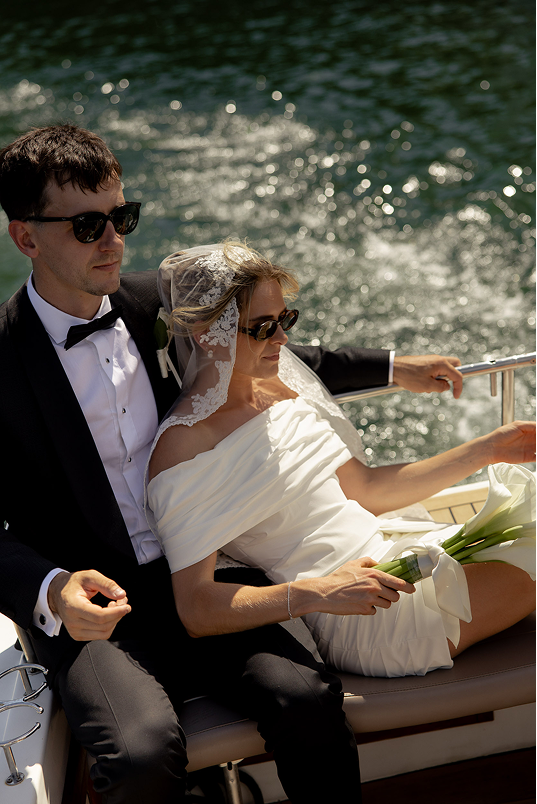 A bride and groom in sunglasses riding on a boat, with the bride holding a bouquet of white calla lilies.