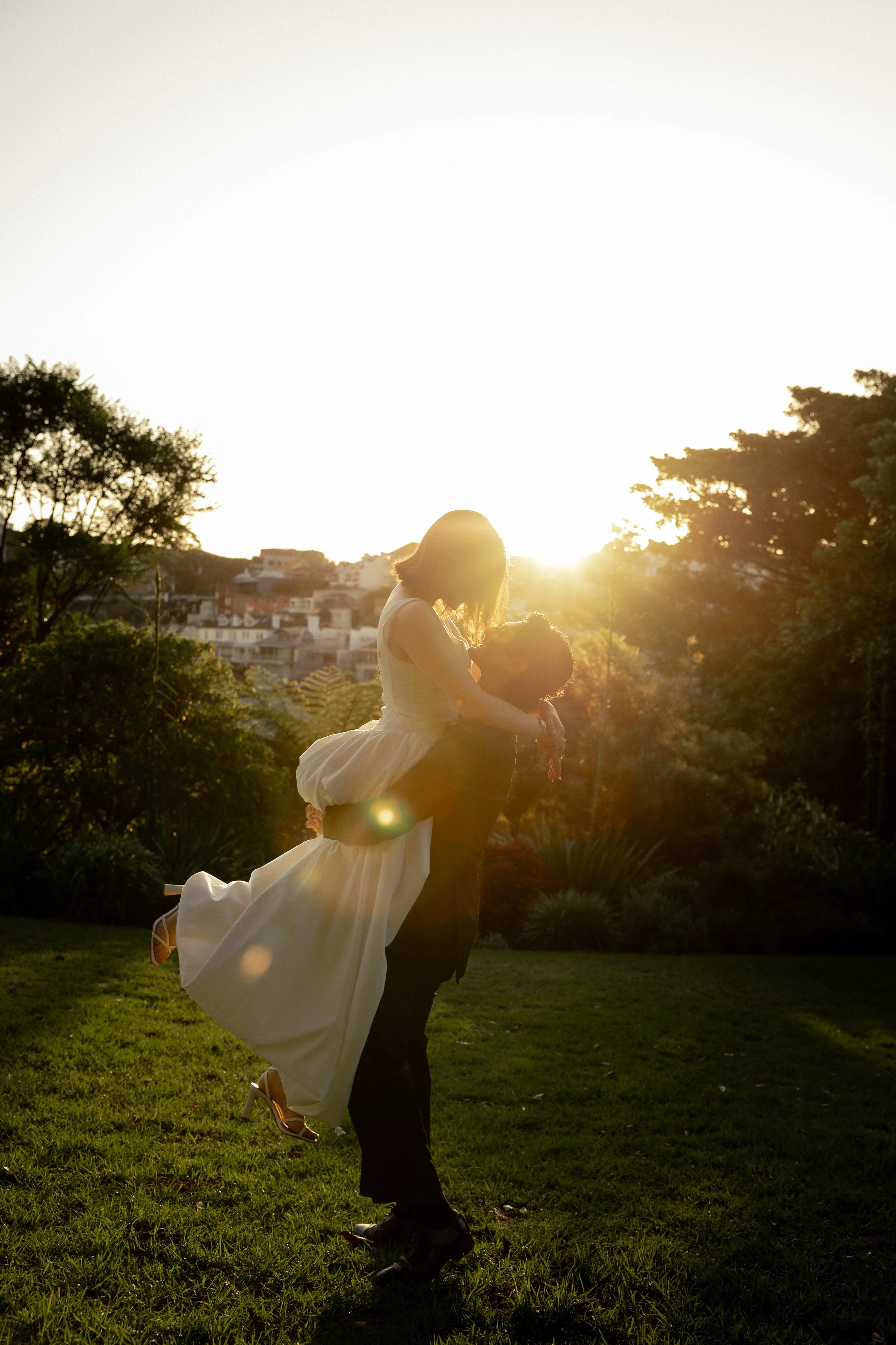 A couple at sunset, with the woman in a white dress being lifted by the man in a dark suit, outdoors with trees and buildings in the background.
