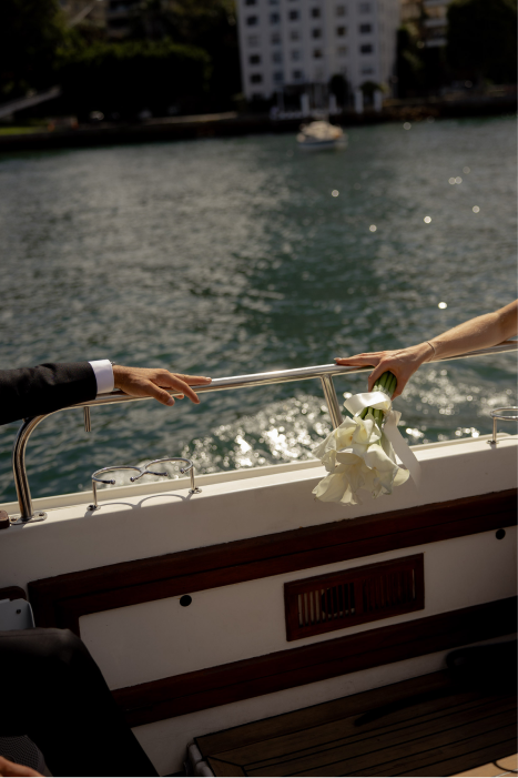 Two people exchanging a bouquet of white calla lilies on a boat with water and city buildings in the background.