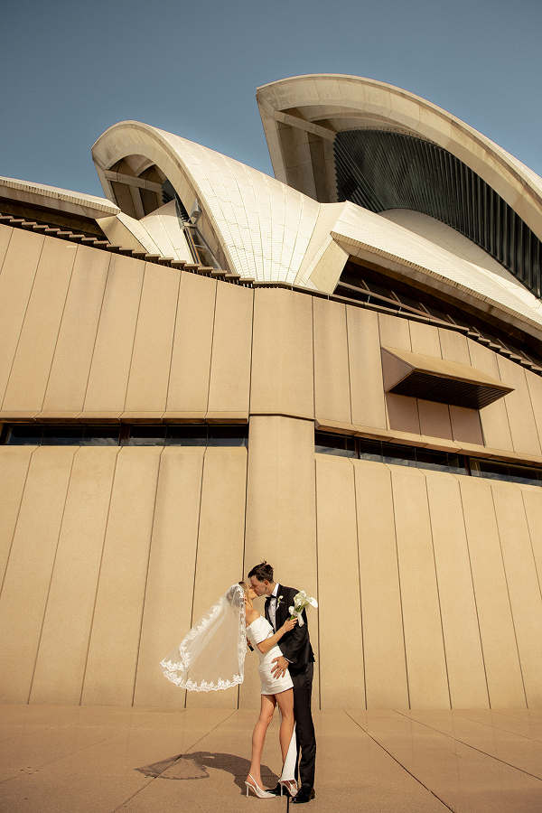 A bride and groom kissing in front of the Sydney Opera House, with the modern architectural structure behind them.