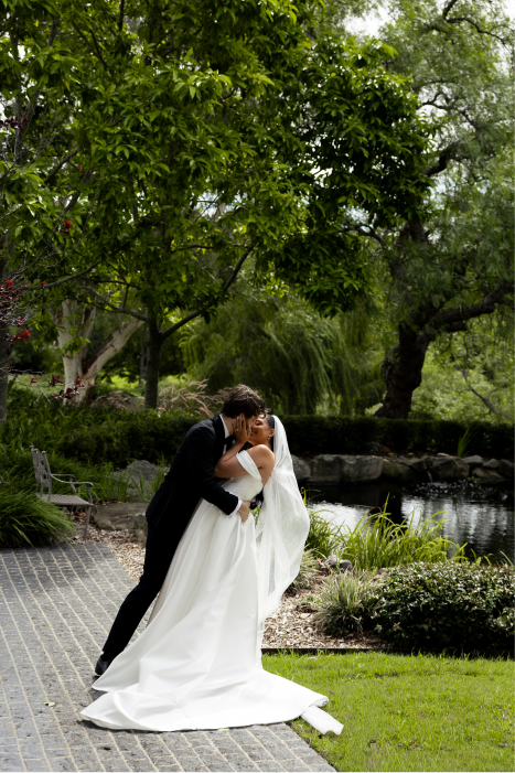 A bride and groom sharing a kiss in a park with lush green trees and a pond.