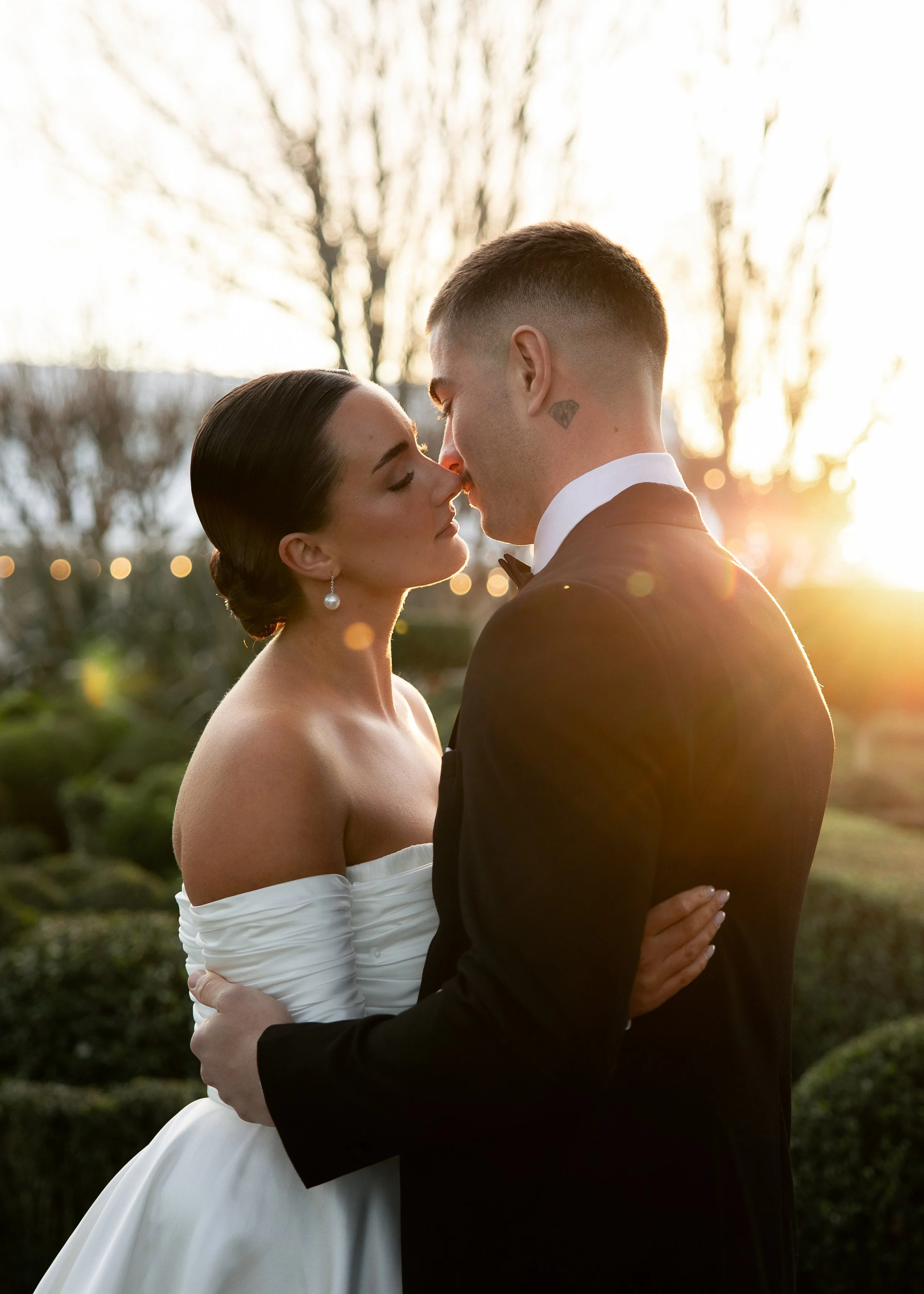 a bride and groom lean in for a kiss with the sunlight behind them at a Sydney wedding venue. the Bride wears a long sleeve white gown and the man wears a black tux, photographed by Haze Photo and Film.