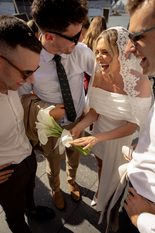 Bride in a white dress with a lace veil holding a bouquet of white calla lilies, laughing with three men in white shirts and dark trousers at an outdoor wedding reception.