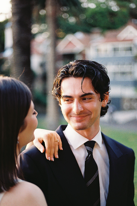 A man and woman outdoors, smiling at each other. The man is wearing a suit and tie, and the woman has her hand on his shoulder.