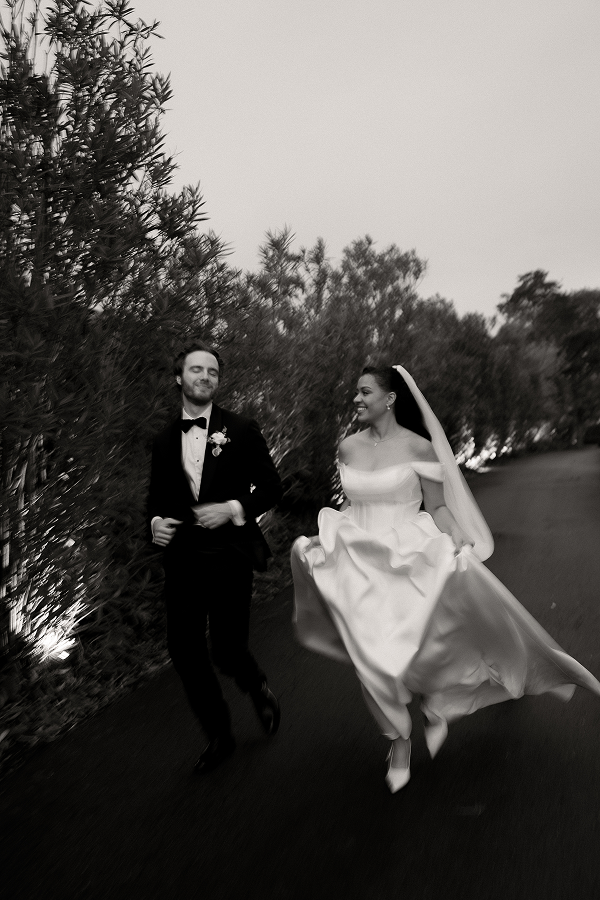 A black and white photo of a bride and groom running outdoors. The groom is wearing a tuxedo with a bow tie and boutonniere. The bride is in a flowing wedding dress with a veil, smiling at the groom. They are on a pathway lined with trees and bushes.