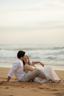 A couple sitting on the beach with the ocean in the background, the man wearing glasses and a white shirt, the woman in a white dress, both relaxing and enjoying the seaside view.