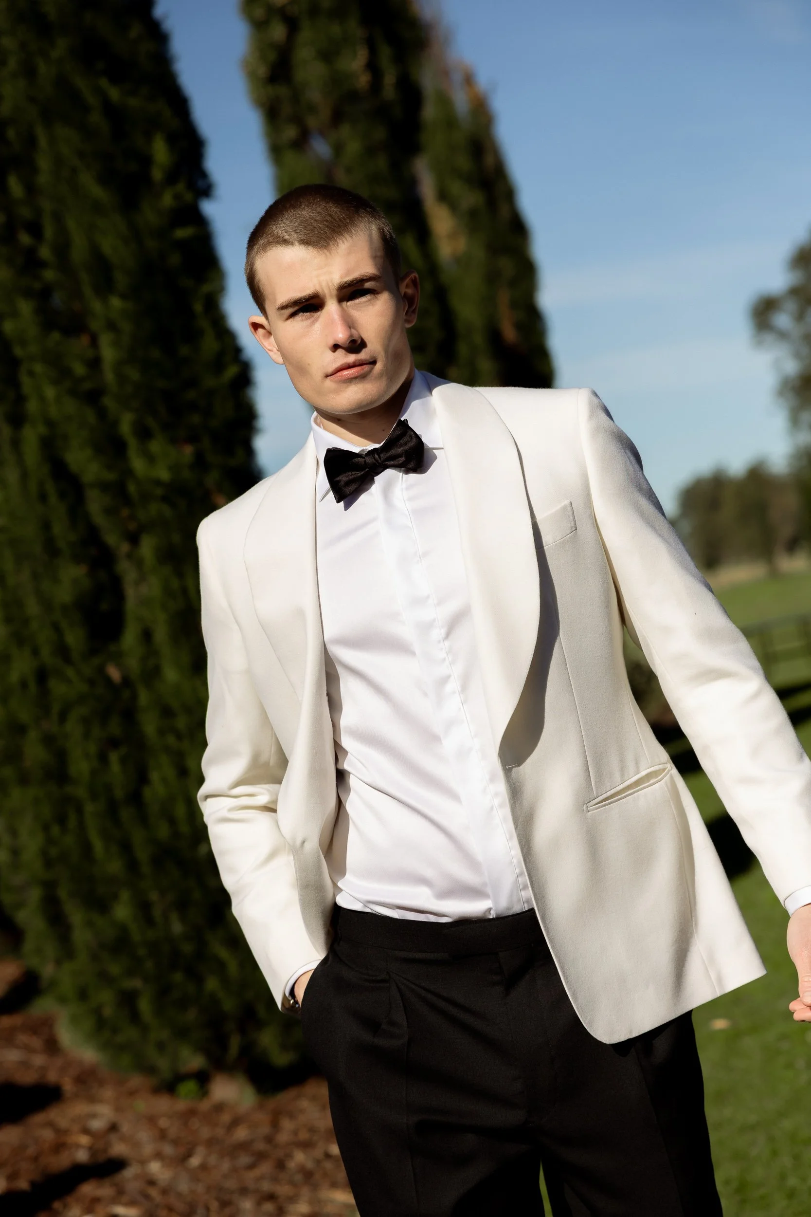 A groom in a white tux walks towards the camera in the morning of his Sydney wedding. The sky is blue and there are tall green tress behind him, photographed by Haze Photo and Film.