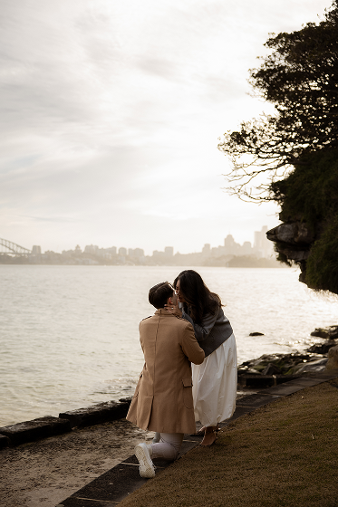 A couple is kissing by the water on a rocky shoreline, with a city skyline and cloudy sky in the background.