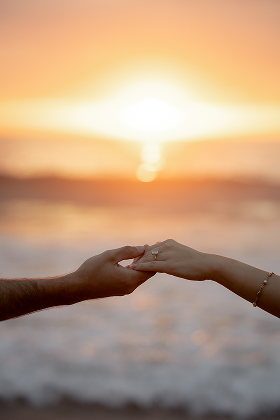 Two hands reaching out and touching in front of a sunset over the ocean.