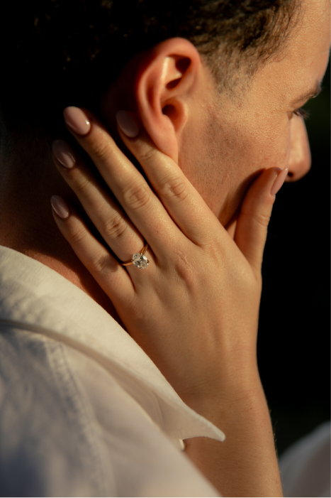 Close-up of a woman's hand with a diamond engagement ring, placed on her neck, with her face partially visible and wearing a light-colored shirt.
