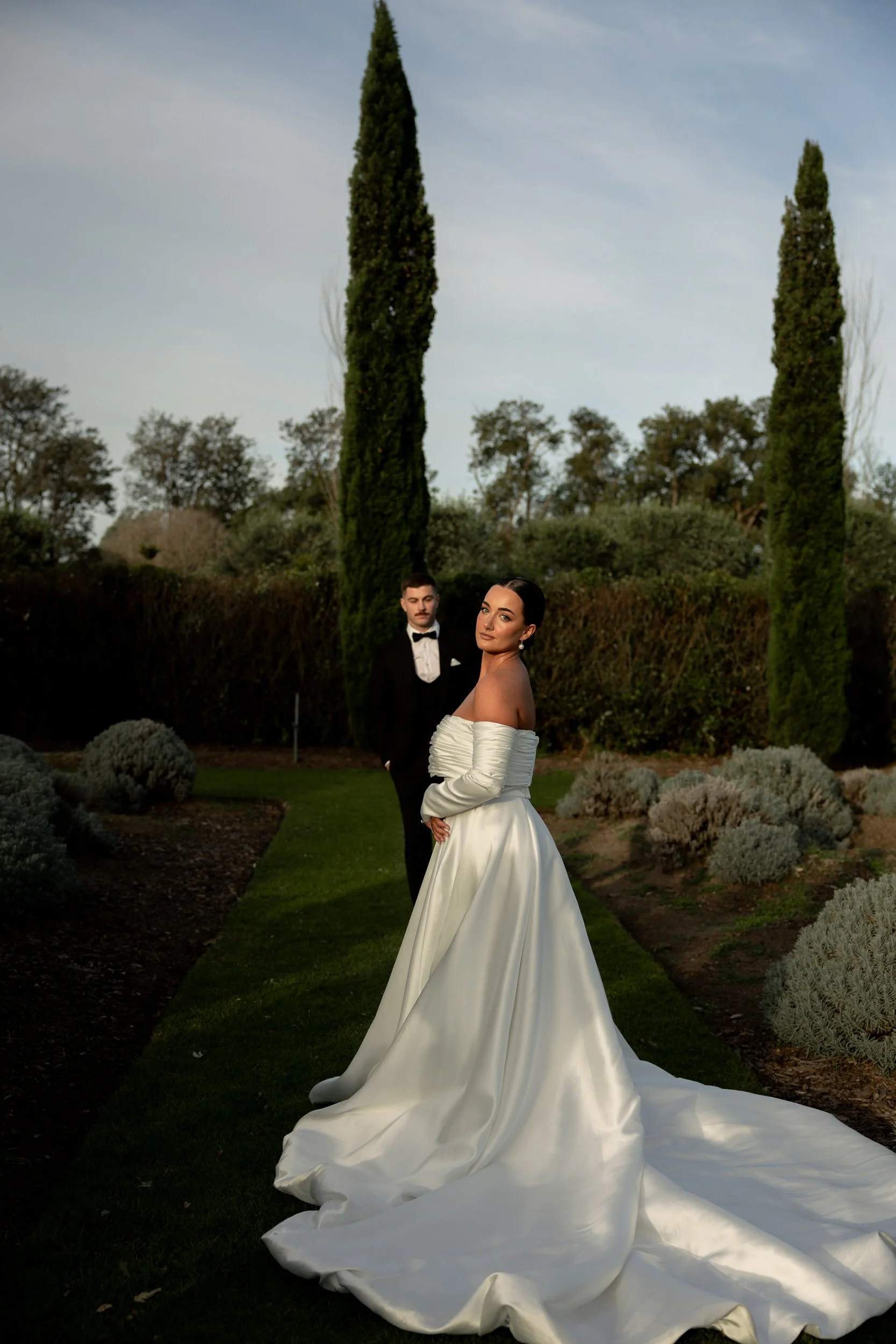 A bride in a white off-the-shoulder wedding gown with a long train stands outdoors in a landscaped garden with tall cypress trees and shrubbery, with a man in a tuxedo in the background.