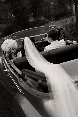 A bride with a veil and bouquet sitting next to a groom in a vintage convertible car.