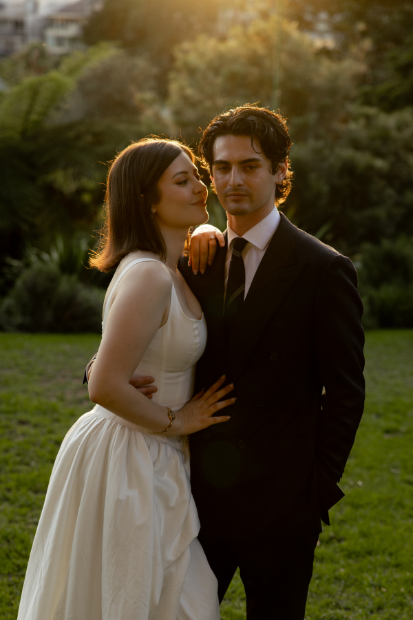 A woman in a white dress and a man in a black suit stand close together outdoors at sunset in a park, with lush greenery and trees in the background.