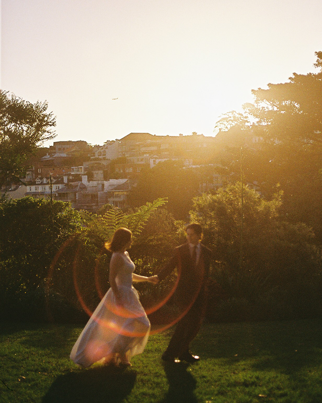 A couple holding hands in a garden during sunset, with a cityscape in the background and lens flare from the sunlight.