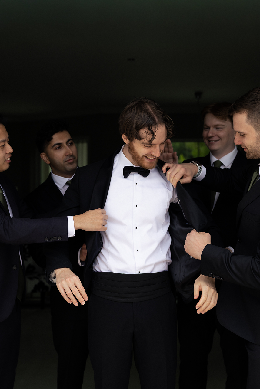 A groom in a tuxedo with a bow tie is surrounded by friends helping him get dressed at a wedding.