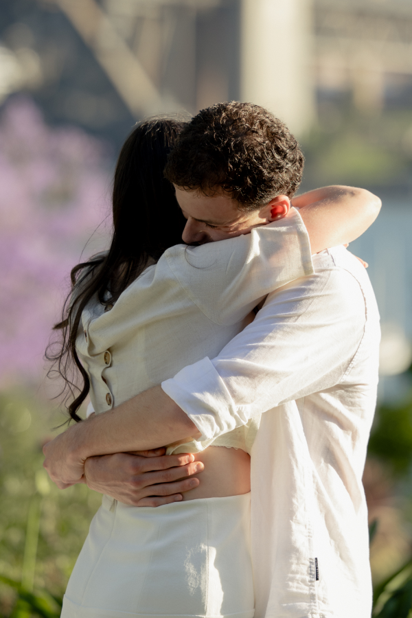 A couple embracing outdoors, with blurred greenery and water in the background.