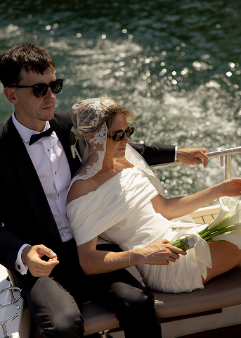 A newlywed couple on a boat ride, with the groom in a tuxedo and the bride in a white dress holding a small bouquet of flowers, sailing on water during daytime.