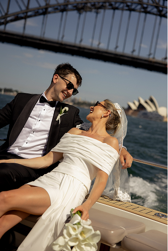 A newlywed couple in wedding attire enjoying a boat ride under the Sydney Harbour Bridge with the Sydney Opera House visible in the background.