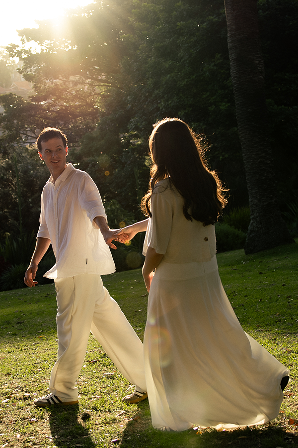A young man and woman holding hands and dancing outdoors in a park during sunset, dressed in light-colored clothing.