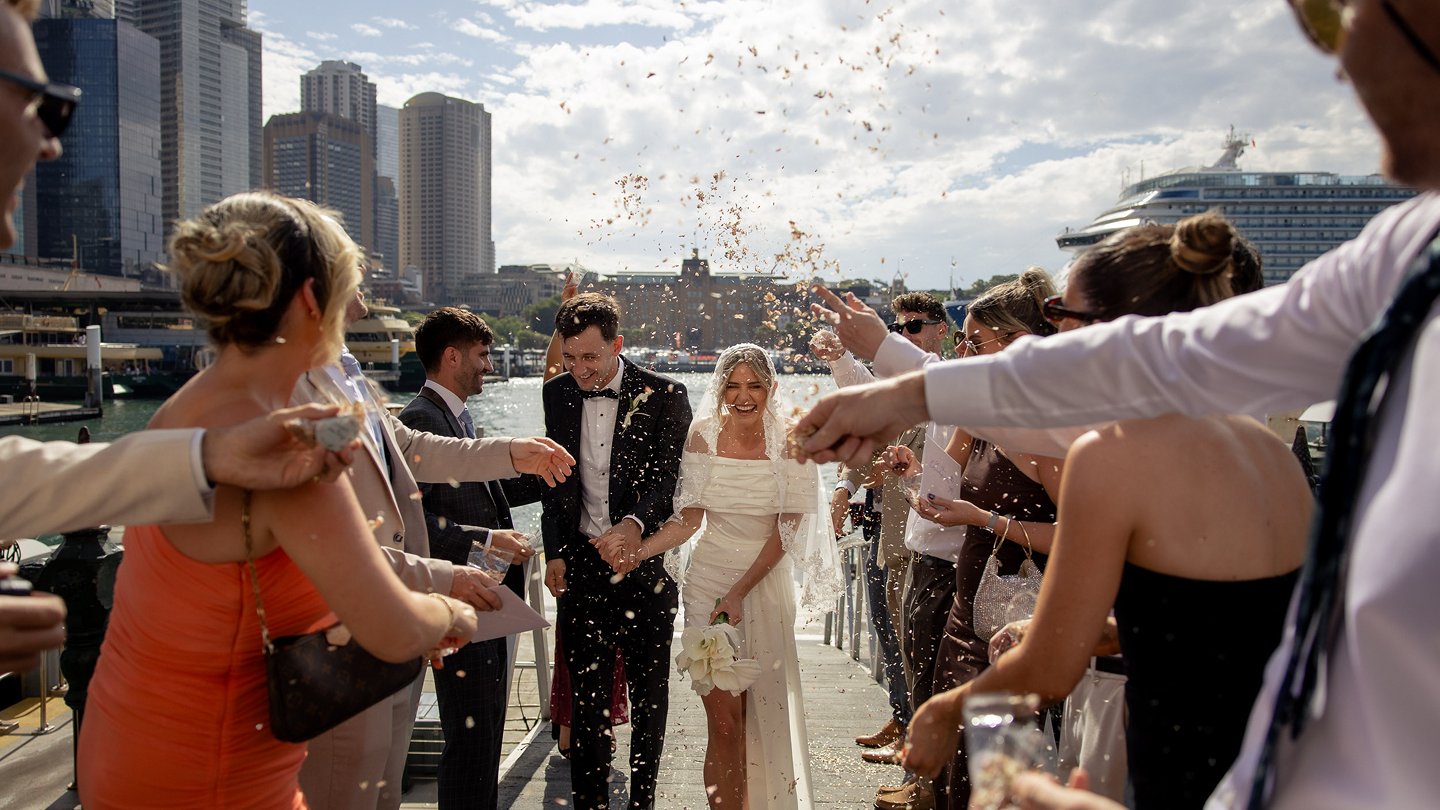 Bride and groom walking on a dock surrounded by friends and family celebrating with confetti in a city skyline background.