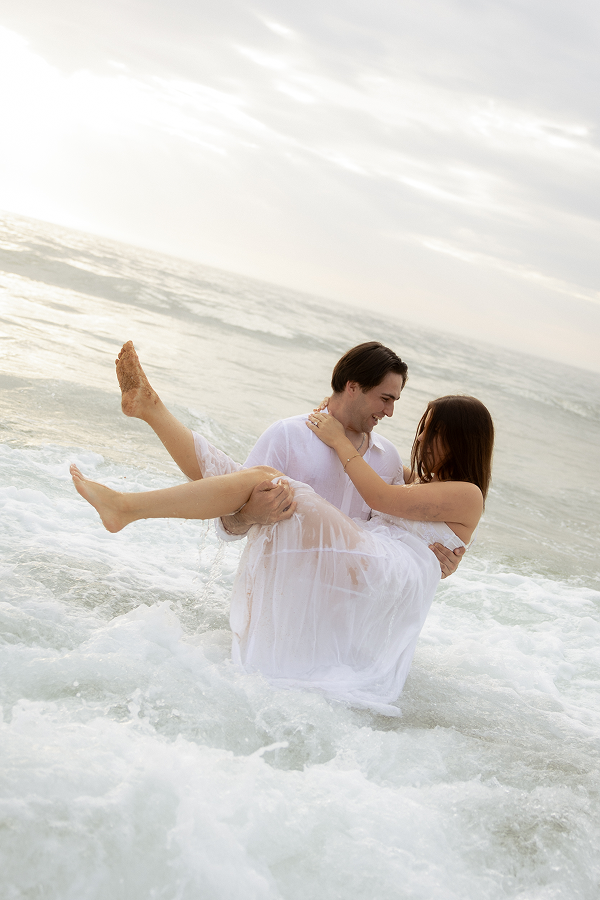 A couple in white clothes enjoying and splashing in the ocean, with the man carrying the woman in his arms near the shoreline.
