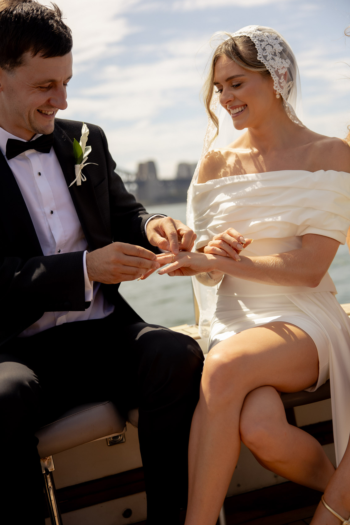 A newlywed couple on a boat, with the groom placing a ring on the bride's finger as they smile at each other against a river and city skyline background.