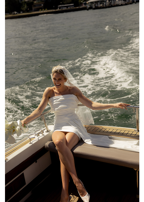 A woman in a white wedding dress sitting on a boat, smiling, with water in the background.