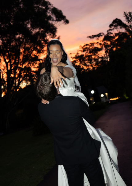 A joyful woman in a wedding dress being lifted by a man in a tuxedo during sunset outdoors.