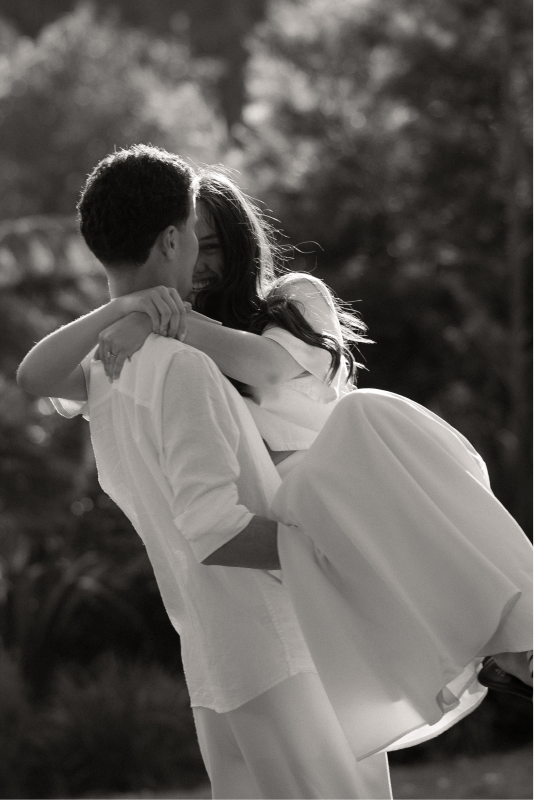 A black and white photo of a couple embracing outdoors, the man holding the woman in his arms and both smiling.