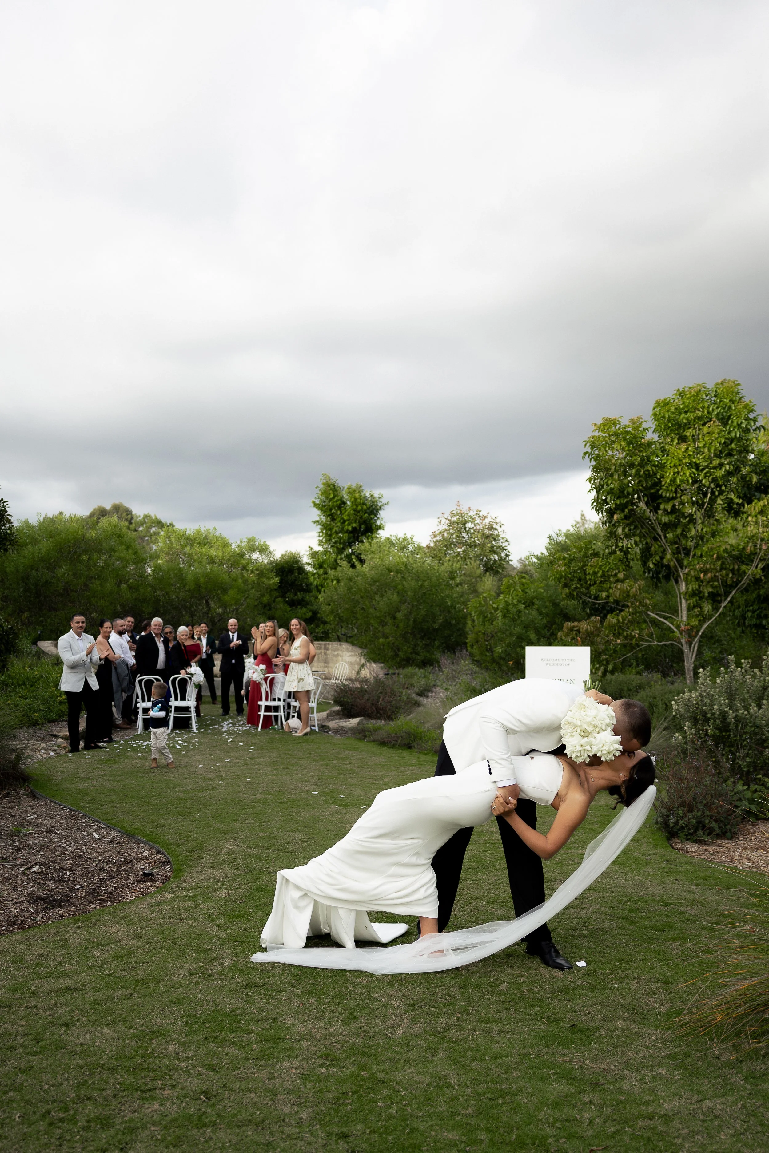 A Bride and Groom do a dip kiss at the end of the aisle with the ceremony party watching on.