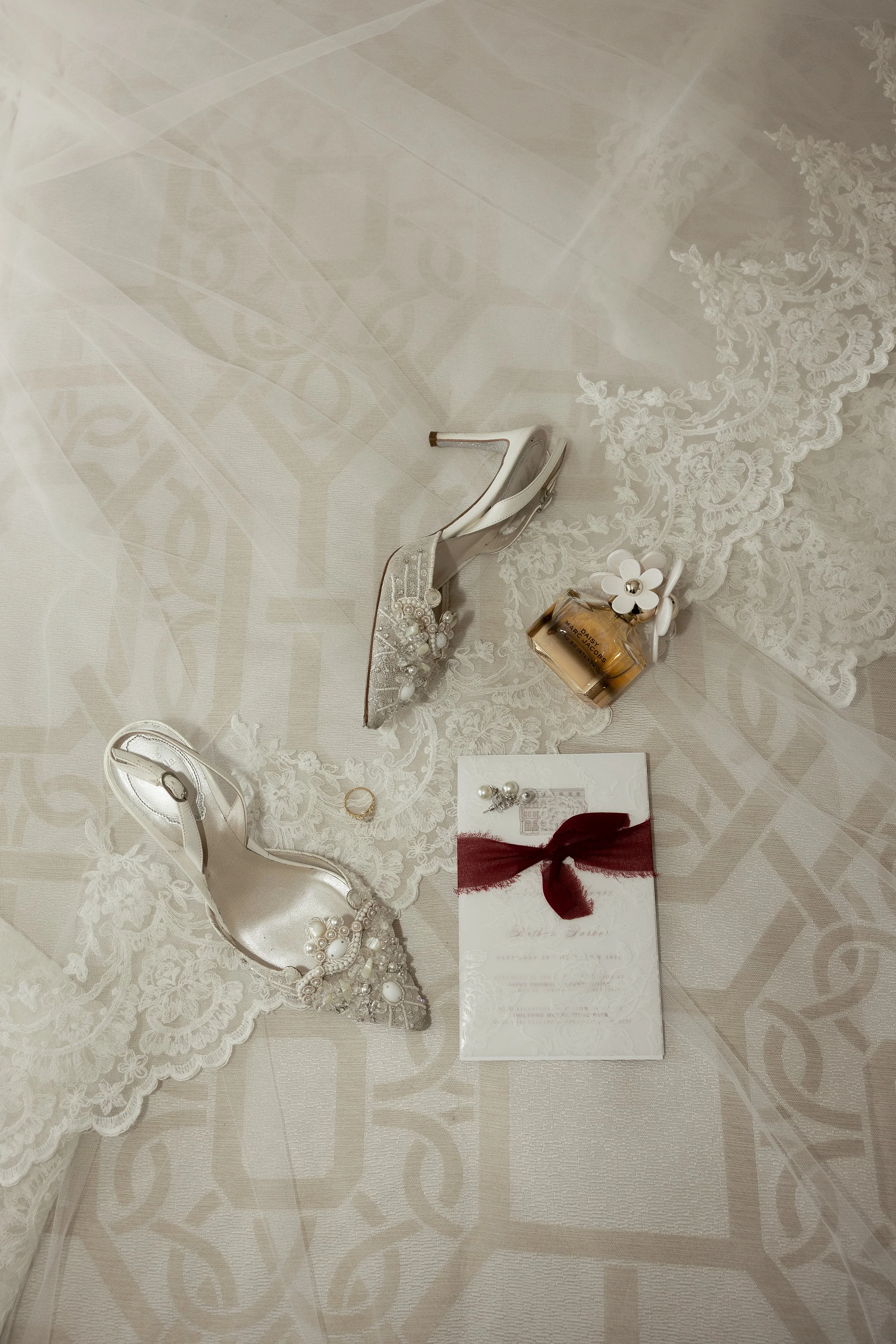 A brides getting ready details. Her white shoes, wedding rings, perfume and wedding invitations lie on a bed on top of her veil, photographed by Haze Photo and Film 